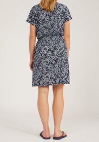 Kobieta stojąca tyłem, ubrana w granatową sukienkę z małym białym wzorem kwiatowym i granatowe klapki na jasnym tle.