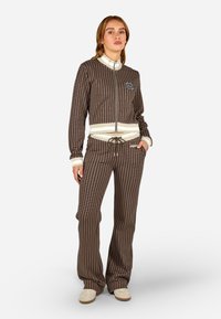 Hnedý vzorovaný teplákový komplet vyrobený z mäkkej látky, ktorý obsahuje zipsovú bundu a nohavice na šnúrku s kontrastnými bielymi akcentmi.