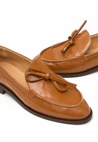 Pár lesklých světle hnědých kožených mokasín s kulatými špičkami a ozdobnými mašlemi na vrchu, s nízkými černými podpatky a prošitými detaily.