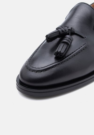 Μαύρο δερμάτινο μοκασίνι με ραφές και δύο διακοσμητικές φούντες στο μπροστινό μέρος, εμφανιζόμενο σε λευκό φόντο.