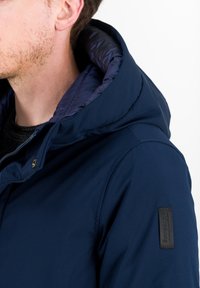 Marineblauwe jas met een gestructureerd exterieur, een glanzende voering van de capuchon, rits-sluiting en een zwart logo-patch op de mouw.