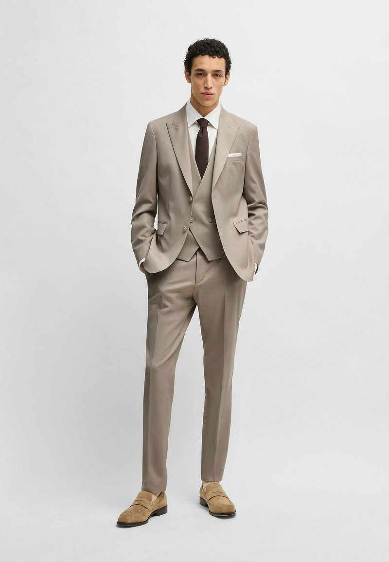 Completo tre pezzi di un leggero beige con blazer a un bottone, pantaloni abbinati e gilet. Completato con una camicia bianca e una cravatta scura.