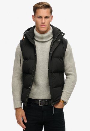 Chaleco de plumas negro con capucha, sobre un suéter de cuello alto acanalado gris claro y jeans oscuros. Presenta accesorios de metal plateado y secciones acolchadas prominentes.