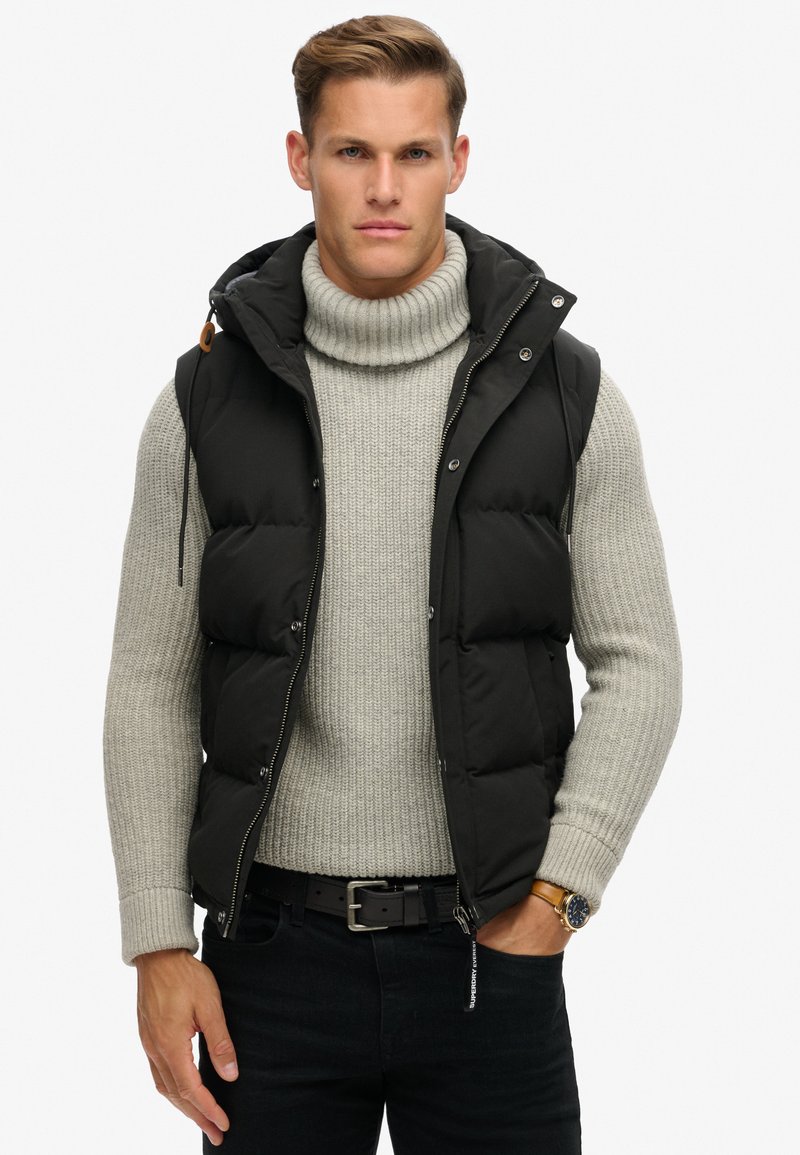 Schwarze Steppweste mit Kapuze über einem hellgrauen, gerippten Rollkragenpullover und dunklen Jeans. Verfügt über silberne Beschläge und auffällige gesteppten Abschnitte.