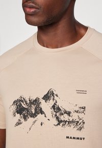 Beżowa koszulka bawełniana z czarną grafiką gór, z okrągłym dekoltem i rękawami raglanowymi. Nazwa marki "Mammut" nadrukowana na dole.