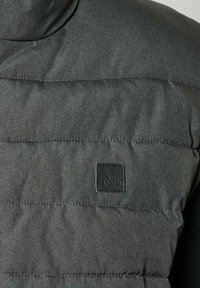 Chaqueta acolchada gris con cuello alto, que presenta un tejido texturizado, costuras horizontales y un parche de logo negro en el pecho.
