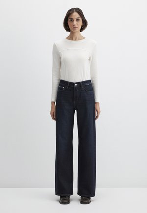 White long-sleeve top with ribbed texture; paired with dark blue wide-leg jeans. Model stands against a plain light background.