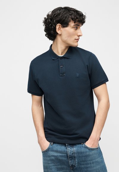 Νεαρός άνδρας με σγουρά μαλλιά που φοράει μπλε πόλο μπλουζάκι και μπλε τζιν παντελόνι, στέκεται με τα χέρια στις τσέπες, κοιτάζοντας προς τα πλάγια.