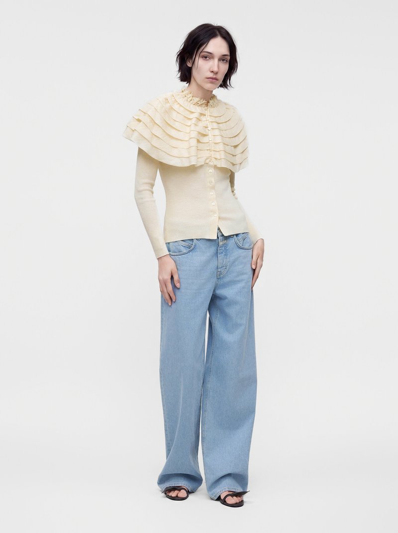 Woman wearing light beige layered ruffled short cape, beige buttoned sweater, wide light blue jeans, and black open-toe sandals standing on white background.