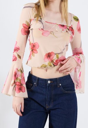 Kobieta nosząca przezroczysty kwiatowy crop top z długimi rękawami dzwonowymi oraz ciemnoniebieskie, z wysokim stanem jeansy, stojąca na jednolitym tle.