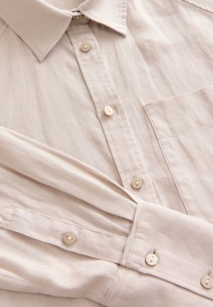 Smėlio spalvos marškiniai su sagomis ir apykakle, pagaminti iš tekstūruotos medžiagos. Yra priekinis kišenė ir perlinių sagelių eilutė palei sagų juostelę bei rankogalius.