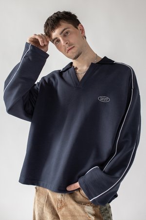 Navy long-sleeve polo shirt with a V-neck, white piping along the sleeves, and a logo patch. Worn with distressed beige pants.