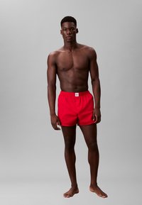 Pantaloni da bagno rossi con vita elastica e apertura anteriore. Il tessuto sembra leggero. Il modello è in piedi a piedi nudi su uno sfondo grigio.