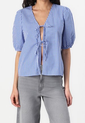 Kvinde iført blåt og hvidt ternet bluse med pufærmer og bindebånd foran, kombineret med lysegrå højtaljede jeans.