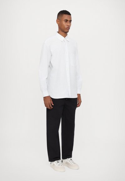 Camisa blanca de manga larga con botones y un bolsillo, con un sutil patrón de rayas verticales. Combinada con pantalones negros de corte relajado y zapatillas.
