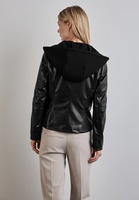 Giacca in faux pelle nera con un grande cappuccio nero e un design aderente. Dettagli delle cuciture e maniche lunghe. Indossata con pantaloni chiari.