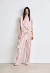 Helle rosa, vertikal gestreifte Bluse mit kurzen Ärmeln und elastischem Taillenbund, kombiniert mit passenden weit geschnittenen Hosen. Eine kleine weiße Handtasche wird gehalten.