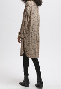 Blouse beige à manches longues avec un motif géométrique noir ; coupe décontractée ; tissu texturé ; fentes sur les côtés ; associée à un pantalon slim noir et des bottines.