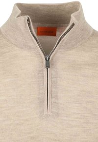 Beige gebreide trui met een halve rits van metaal en een opgerichte kraag, met een oranje merketiket aan de binnenkant van de hals.