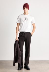 Camiseta de algodão branca com logotipo circular, cinto preto, calças de lã castanhas, usada com um gorro bordeaux e segurando uma jaqueta escura. Sapatos pretos.