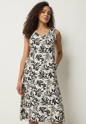 Frau mit lockigem Haar, die ein ärmelloses, knieumspielendes Kleid mit schwarzem Blumenmuster auf weißem Hintergrund trägt und vor einer schlichten Wand steht.