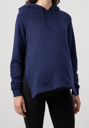 Model draagt een marineblauwe hoodie met een ontspannen pasvorm, gecombineerd met zwarte leggings, staande tegen een effen lichte achtergrond.