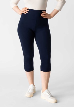 Donkerblauwe capri leggings van rekbaar materiaal met een hoge tailleband. Gecombineerd met witte sneakers met een gestructureerde zool en een gevoerde tong.