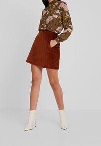 Blouse florale marron à col haut, associée à une jupe évasée en velours côtelé rouille avec poches ; le modèle porte des bottines blanches.