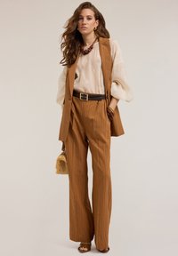 Bruine gestreepte wijde broek gecombineerd met een crèmekleurige blouse met geborduurde details. Geaccentueerd met een blauwe riem en accessorized met een geweven tas.