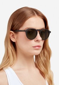 Guľaté čierne slnečné okuliare s tmavými tónovanými sklami. Majú výrazné geometrické rámy a nenápadné označenie na stranici. Hladký matný povrch.