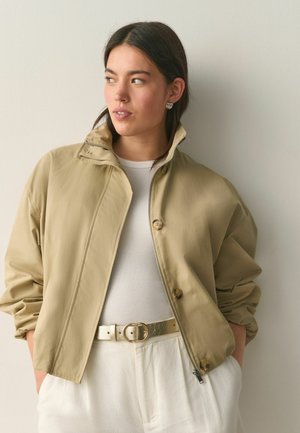 Kobieta ubrana w beżową kurtkę na białej prążkowanej bluzce i białych spodniach z metalicznym złotym paskiem, patrząca na bok, z rękami w kieszeniach.