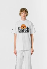 Camiseta blanca de manga corta con un gráfico de un perrito marrón esponjoso y un texto bold negro que dice "Black" con letras adicionales debajo.