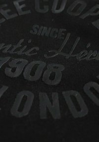 Tissu noir avec texte noir en relief et brillant. Présente les inscriptions "LEE COOPER", "SINCE 1908" et "LONDON" dans différentes polices et tailles.