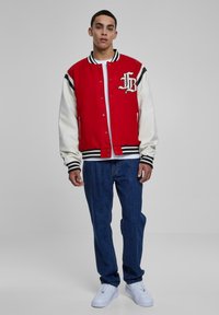 Rød og hvid varsityjakke med sorte detaljer, broderet logo på brystet, og parret med blå jeans og hvide sneakers. Casual pasform.