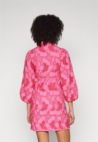 Vestido de manga curta em rosa brilhante com um intricado padrão de renda floral, apresentando mangas volumosas e um colarinho alto, com um acabamento texturizado.