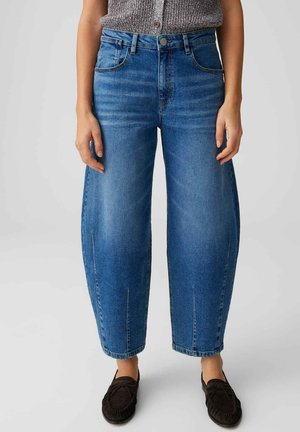 Vrouw die hoge, loszittende blauwe spijkerbroek met voorzakken en zwarte instap-mocassins draagt, staand tegen een effen achtergrond.