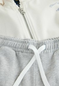 Witte zip-up hoodie met een zilveren rits en geborduurde tekst. In combinatie met grijze sweatpants met een witte koord. Zachte katoenmix.