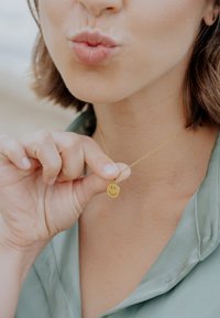 Złoty naszyjnik z okrągłą zawieszką z uśmiechniętą twarzą, zawieszony na delikatnym łańcuszku, noszony na jasnopiętrowej bluzce.
