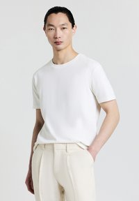 T-shirt bianco a maniche corte con scollatura rotonda, caratterizzato da un tessuto testurizzato. Abbinato a pantaloni beige chiaro, con le mani infilate nelle tasche.