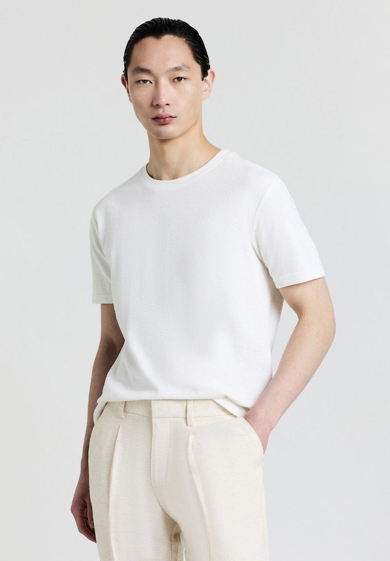 T-shirt bianco a maniche corte con scollatura rotonda, caratterizzato da un tessuto testurizzato. Abbinato a pantaloni beige chiaro, con le mani infilate nelle tasche.