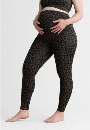 Schwangere Person trägt schwarze und dunkelgrüne Leggings mit Leopardenmuster und Sport-BH, die Hände halten sanft den Bauch, barfuß auf weißem Boden.