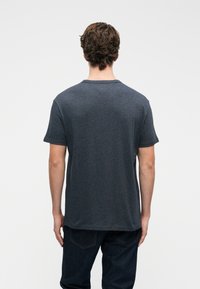 Muž s kvačitými hnědými vlasy, oblečený v tmavě šedém tričku s krátkým rukávem a tmavých džínách, stojící zády k jednoduchému pozadí.