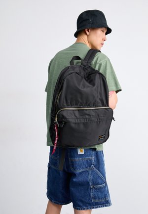 Mochila negra con textura suave, bolsillo frontal con cremallera y herrajes metálicos. Cuenta con correas ajustables y una etiqueta de marca. Llevada por una persona.