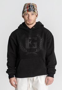 Sudadera con capucha de felpa negra, con un gran logotipo en relieve y un diseño de laurel, que cuenta con un bolsillo frontal y puños acanalados. Se combina con un gorro de punto a cuadros.