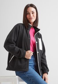 Black fleece jacket with white accents, featuring a snap closure, high collar, and front pocket. Worn over a pink shirt and blue jeans.