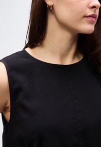 Γυναίκα που φοράει μαύρο αμάνικο τοπ και χρυσούς κρίκους σκουλαρίκια, με μακριά καστανά μαλλιά, μπροστά σε απλό ανοιχτό φόντο.