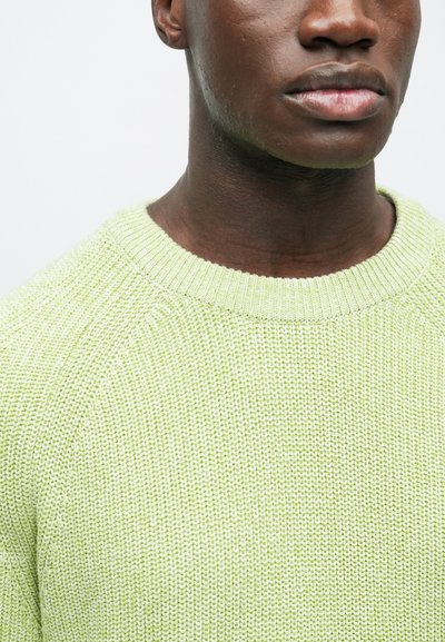Detailný záber na spodnú časť tváre a krk osoby, ktorá má oblečený textúrovaný svetlozelený pletený sveter s okrúhlym golierom.