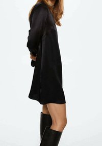 Schwarzes Satin-Kleid mit langen Ärmeln und lässigem Schnitt, das eine glatte Textur und eine knielange Länge aufweist. Getragen mit knielangen schwarzen Stiefeln.