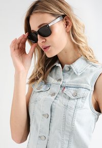 Occhiali da sole neri con un accento metallico argento sulle astine. Il modello indossa un gilet di denim azzurro con bottoni a pressione.