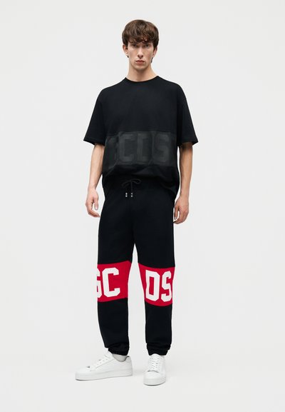 Νεαρός άνδρας που φοράει μαύρο φαρδύ μπλουζάκι και μαύρο παντελόνι με κόκκινες επιφάνειες και λευκά γράμματα, στέκεται μπροστά σε ένα απλό φωτεινό φόντο.